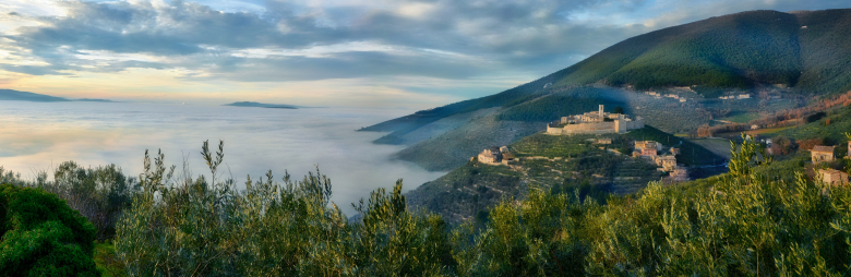 Immagine: Borgo fortificato su una collina circondato dalla nebbia, con case in pietra, mura medievali e uliveti che emergono tra le nuvole 